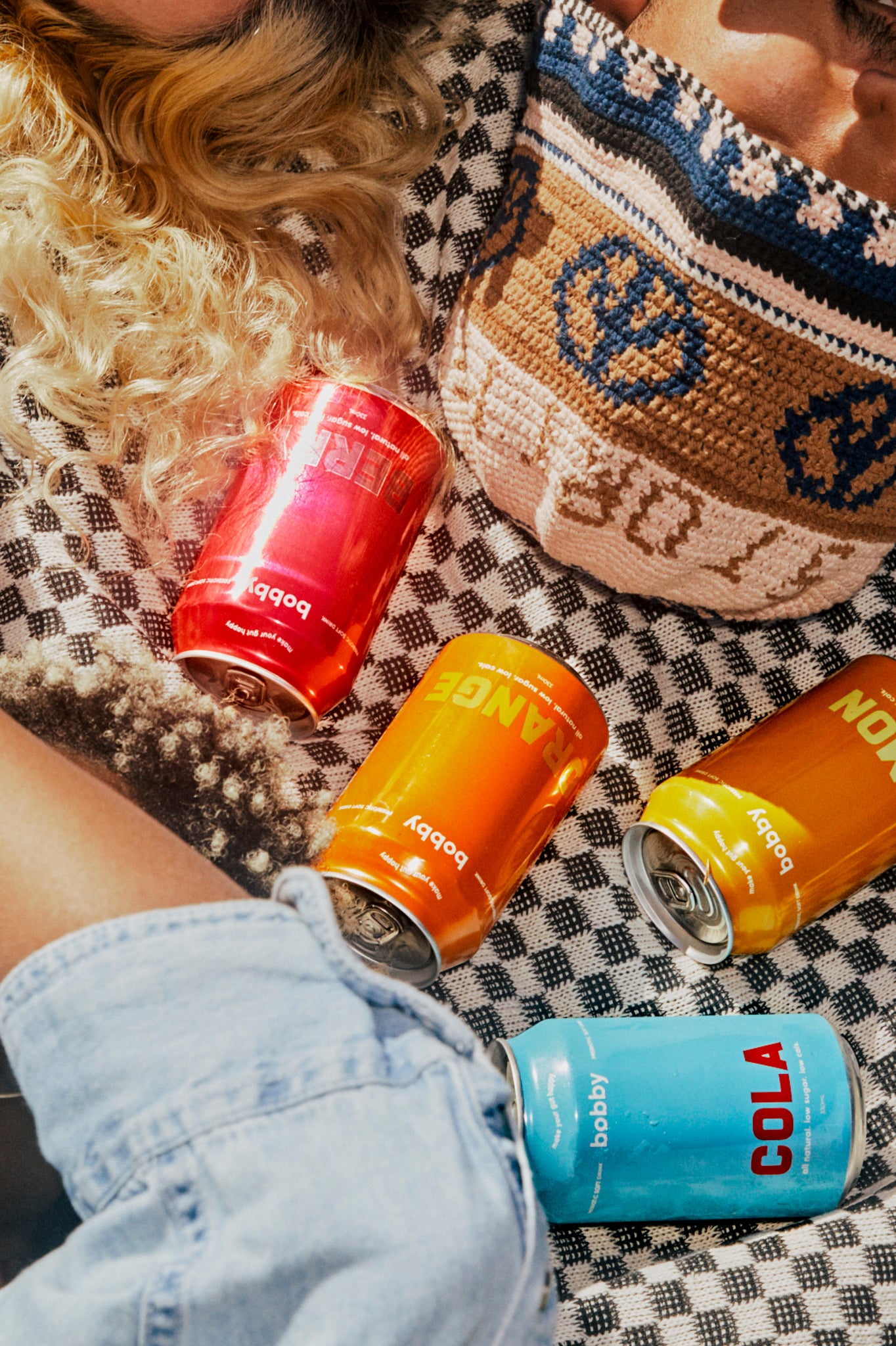 photography by Charlie Hawks, three bobby soda cans, cola, orange, lemon and berry, on a picnic blanket 